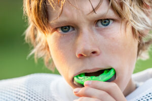Child wearing green sports mouth guard with blue eyes against green background.