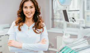Smiling female dentist with arms crossed in modern dental office treatment room.
