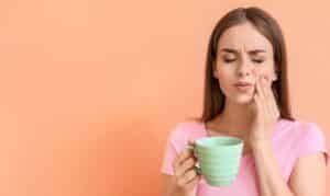 Woman with tooth sensitivity holding green cup and touching her cheek in pain.