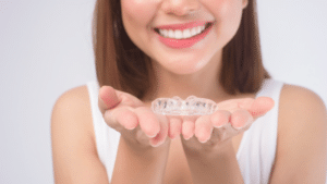 Woman holding clear Invisalign aligner tray, smiling at camera in dental office setting.
