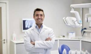 Smiling dentist in white coat standing in modern dental office with treatment chair and equipment.
