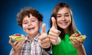 Two happy kids holding healthy sandwiches and giving thumbs up against blue background.