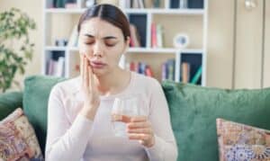Woman holding glass of water while touching her cheek in pain, experiencing tooth discomfort.