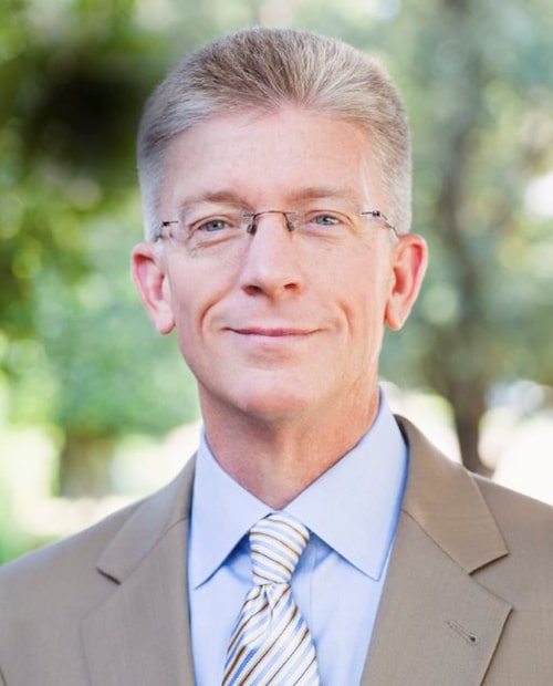 Professional headshot of a man wearing glasses and business attire against a blurred outdoor background.