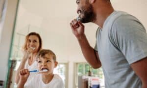 Father and son brushing teeth together in bathroom with mother smiling in background.