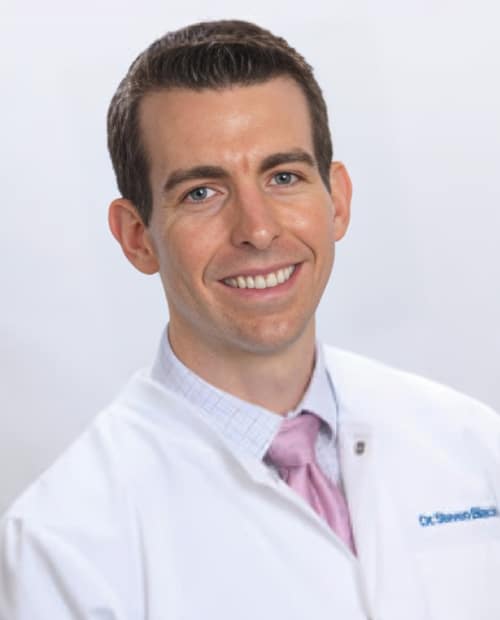 Professional headshot of male dentist in white coat and pink tie smiling at camera