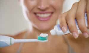 Woman applying toothpaste to electric toothbrush while smiling at home.