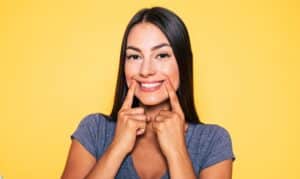 Woman smiling brightly while pointing to her teeth against a yellow background.