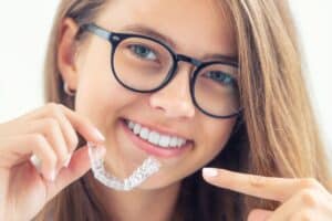 Young woman wearing glasses holds clear Invisalign aligner and smiles brightly.
