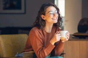 Smiling woman wearing glasses and brown sweater holds coffee cup in dental office.