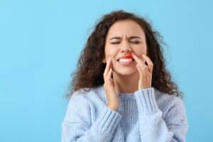 Woman with gum discomfort touching her cheek and smiling against blue background.