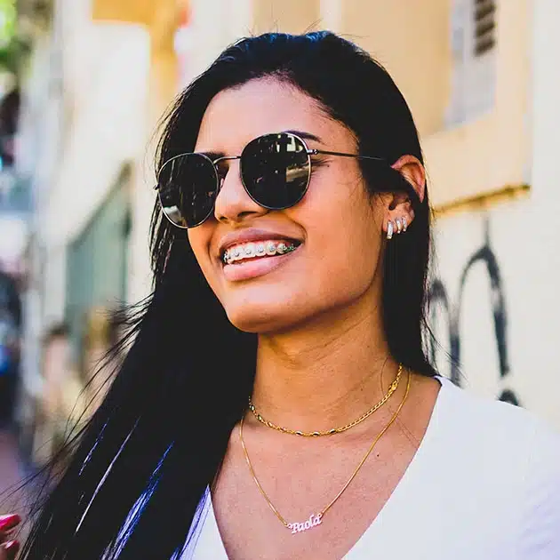 Smiling woman wearing sunglasses and necklace outdoors in urban setting.