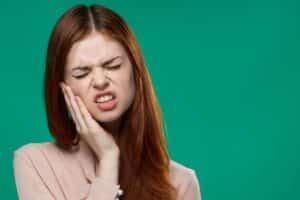 Woman holding her cheek in pain from a cracked tooth, grimacing against a green background.