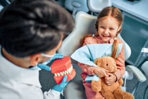 Young girl smiling during pediatric dental visit while holding stuffed bear and examining dental model.